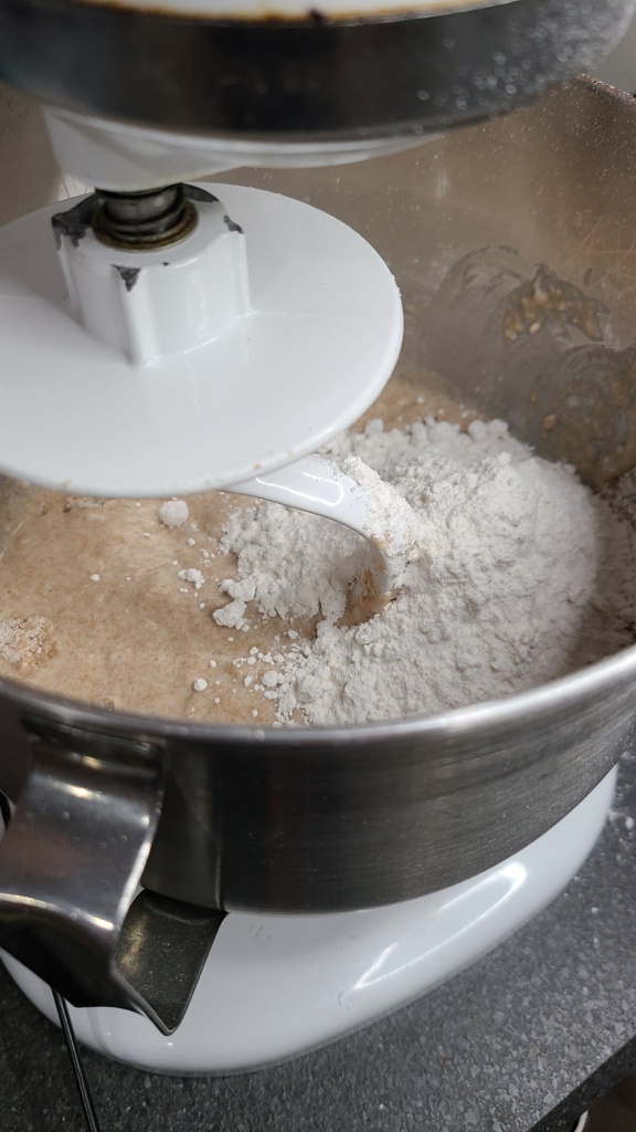 A bowl of a KitchenAid mixer. Inside is bread dough, with all purpose flour being mixed in.