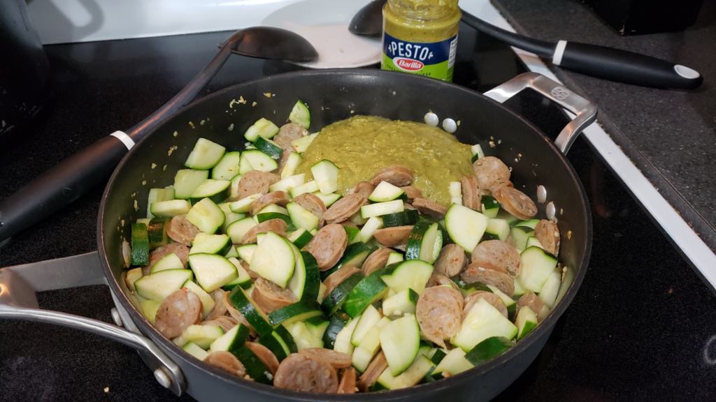 A large skillet with chicken sausage, zucchini, and pesto sauce