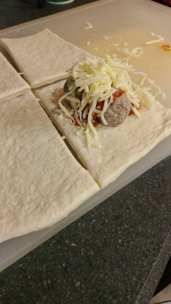 A piece of pizza dough on a cutting board, with meatballs, marinara sauce, and shredded mozzarella on top