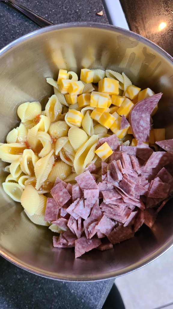 A bowl filled with shell pasta, cheese, salami, and Italian dressing