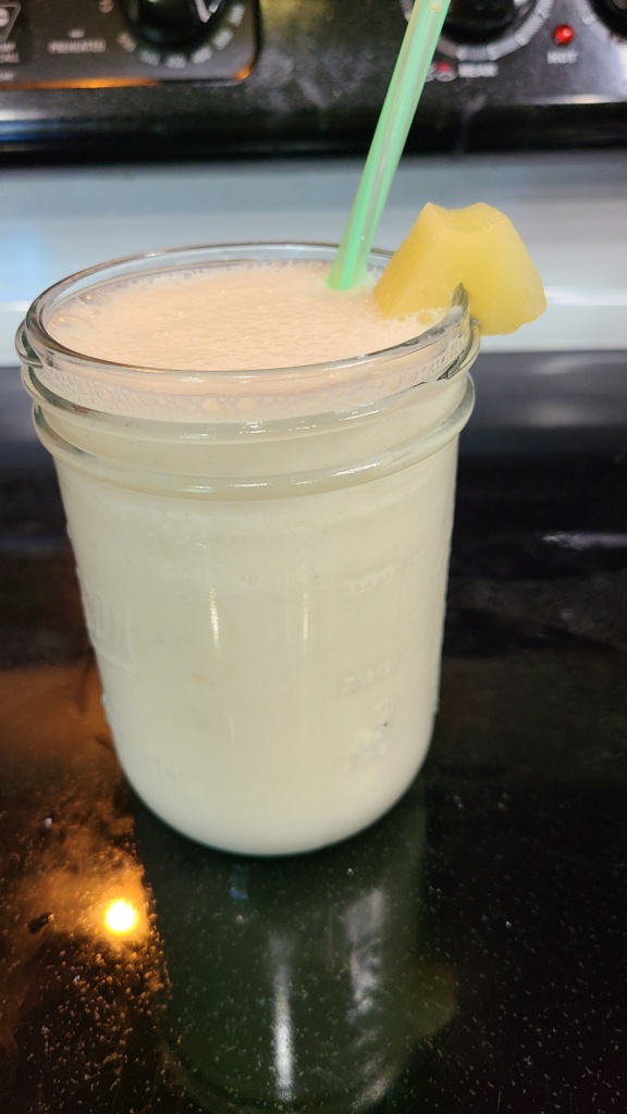 A Mason jar filled with pineapple smoothie, with a straw and a pineapple chunk on the side of the jar