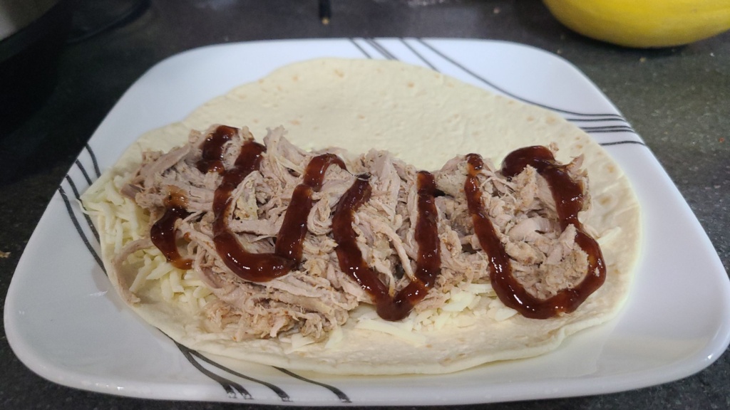 A tortilla with pulled pork, shredded mozzarella, and BBQ sauce on the bottom half