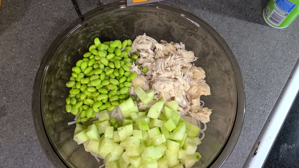 A large bowl filled with rice noodles, chicken, edamame, and cucumber.